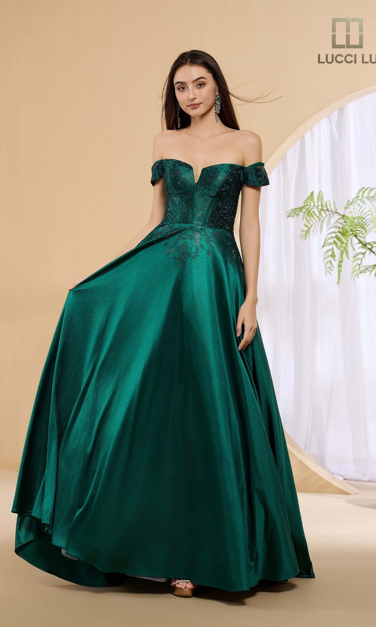 A woman in the Lucci Lu 1483 long prom dress in emerald, featuring an off-shoulder corset bodice and full skirt, stands before a beige backdrop with a circular window and plant.